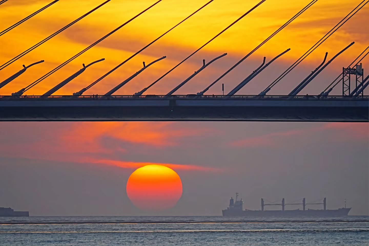 夕陽緩緩沉入海平面，金黃與橘紅的餘暉將天空染得絢麗奪目。壯麗的斜張橋線條交織在晚霞中，遠方的大船伴隨著蛋黃般的落日，勾勒出一幅療癒又詩意的海港黃昏美景 